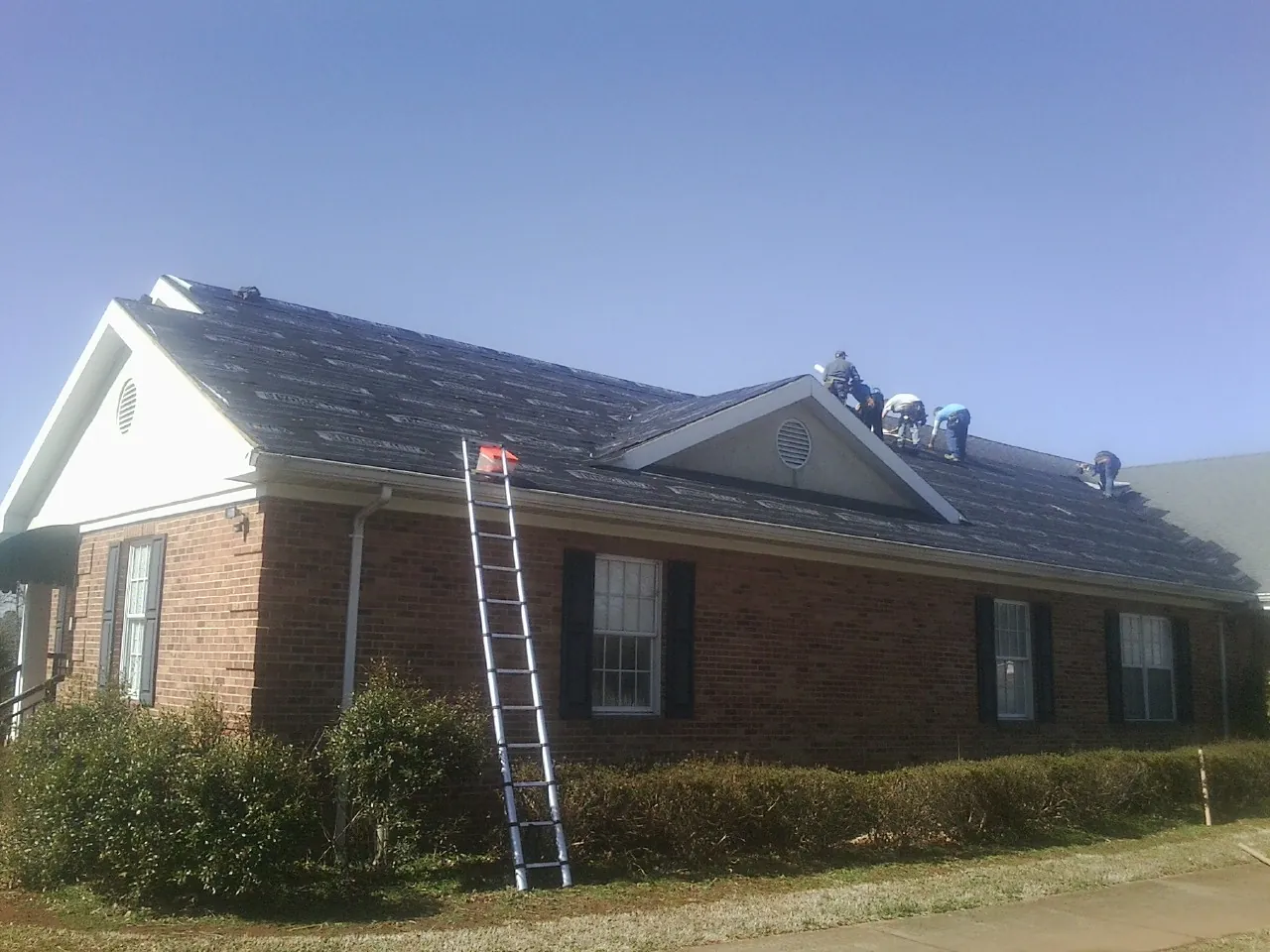 Crew tearing shingles off a brick ranch roof with a ladder set — Metal Roof Repair work by our crew in Mason City